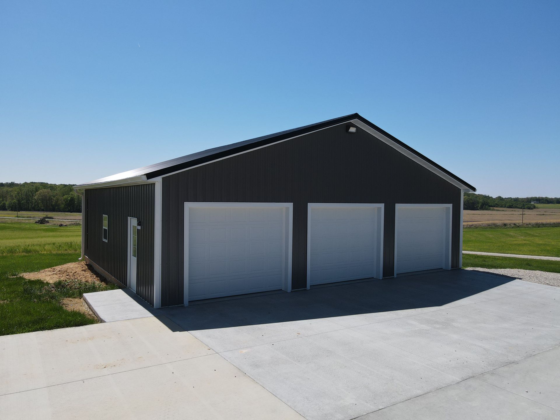 Black three-bay garage with white doors on a concrete driveway in a rural setting