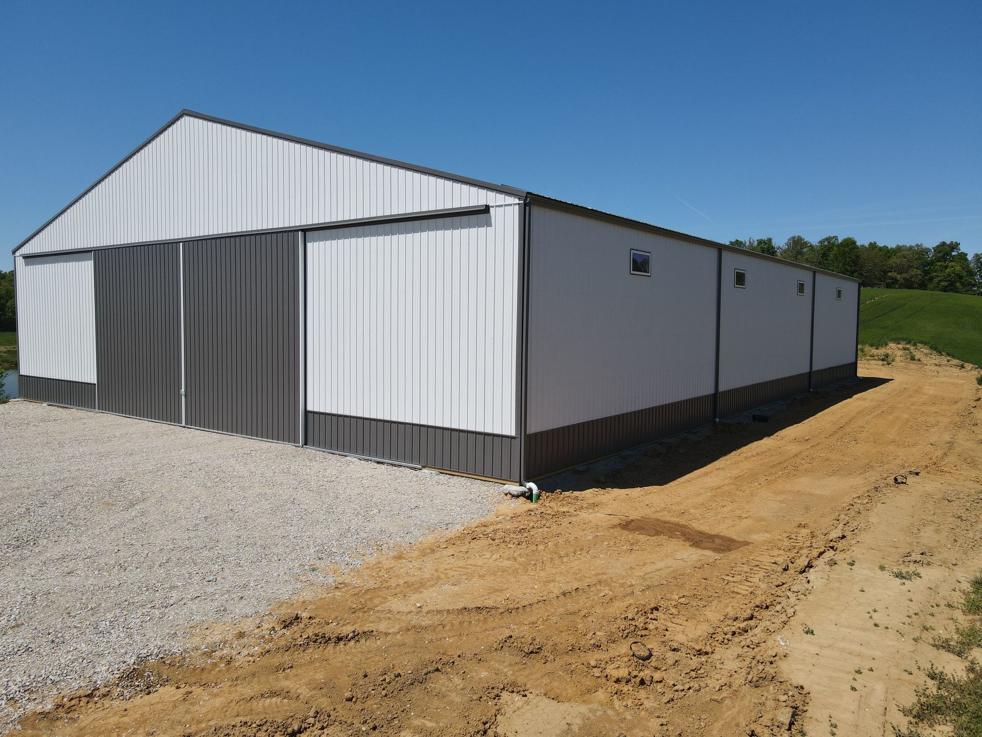 White metal barn-style building beside a gravel lot and dirt road under a clear blue sky