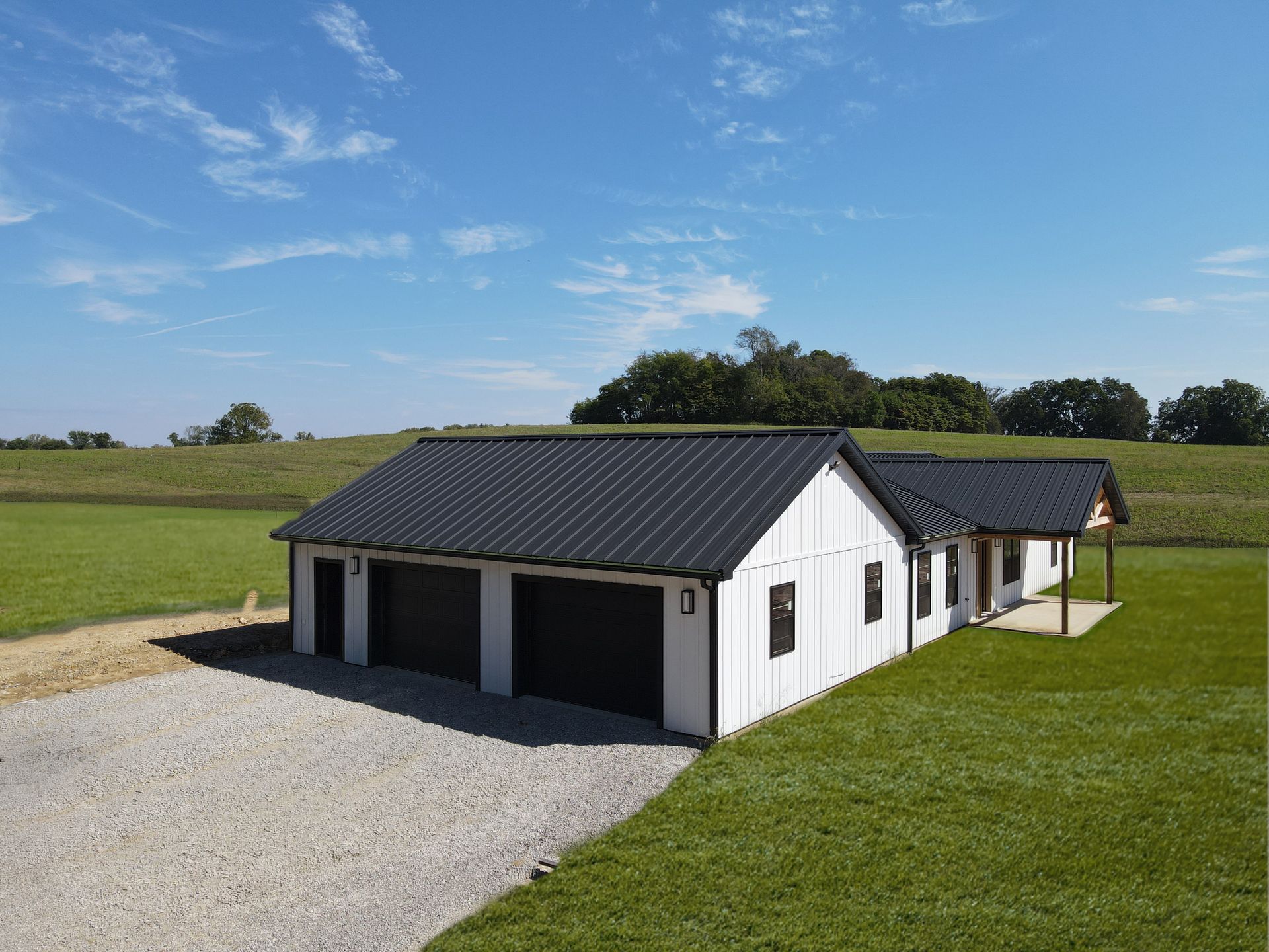 White detached garage with dark roof and gravel driveway in a green rural field under blue sky