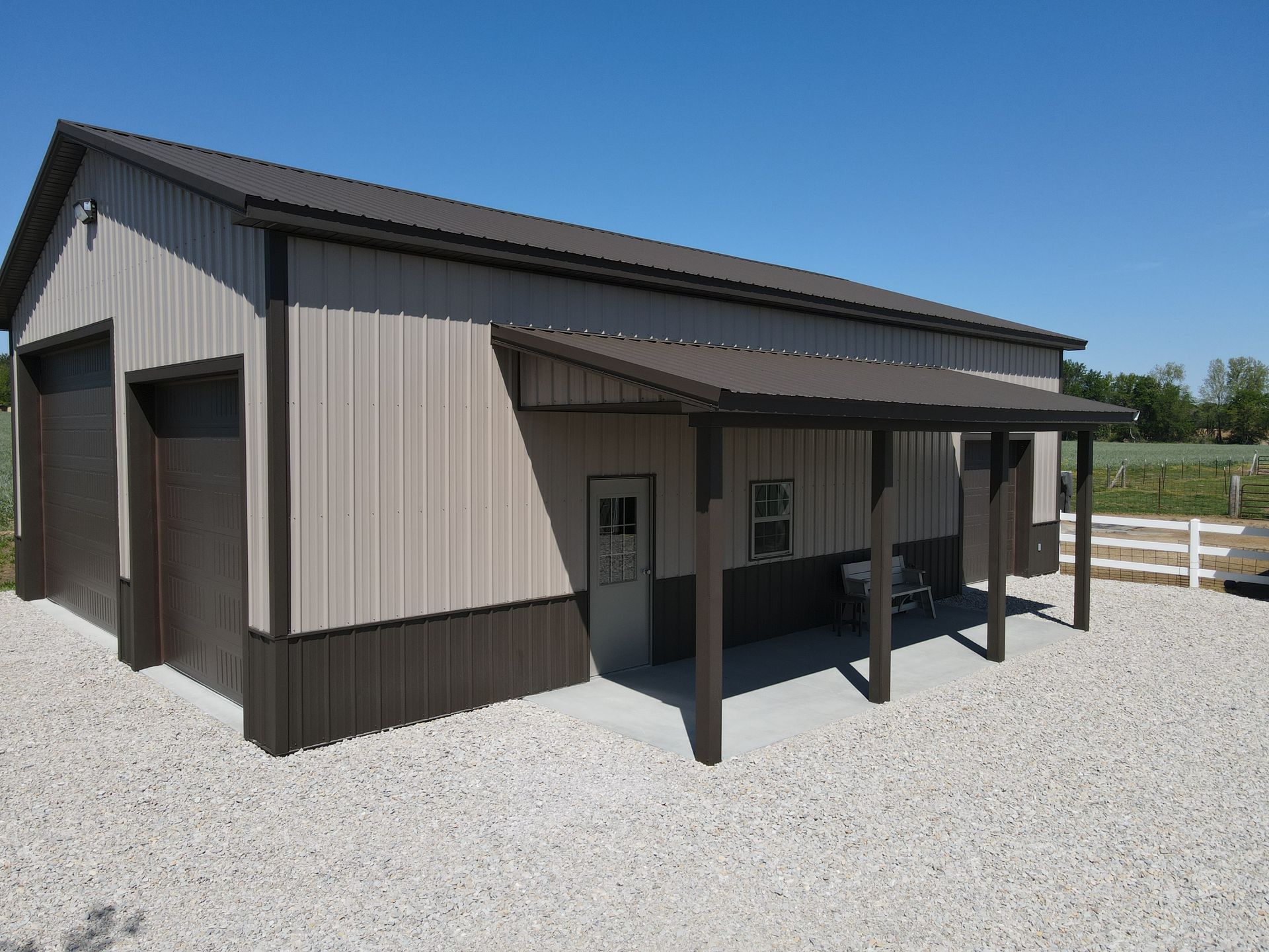 Small tan-and-brown metal barn with a covered porch on a gravel lot under a blue sky