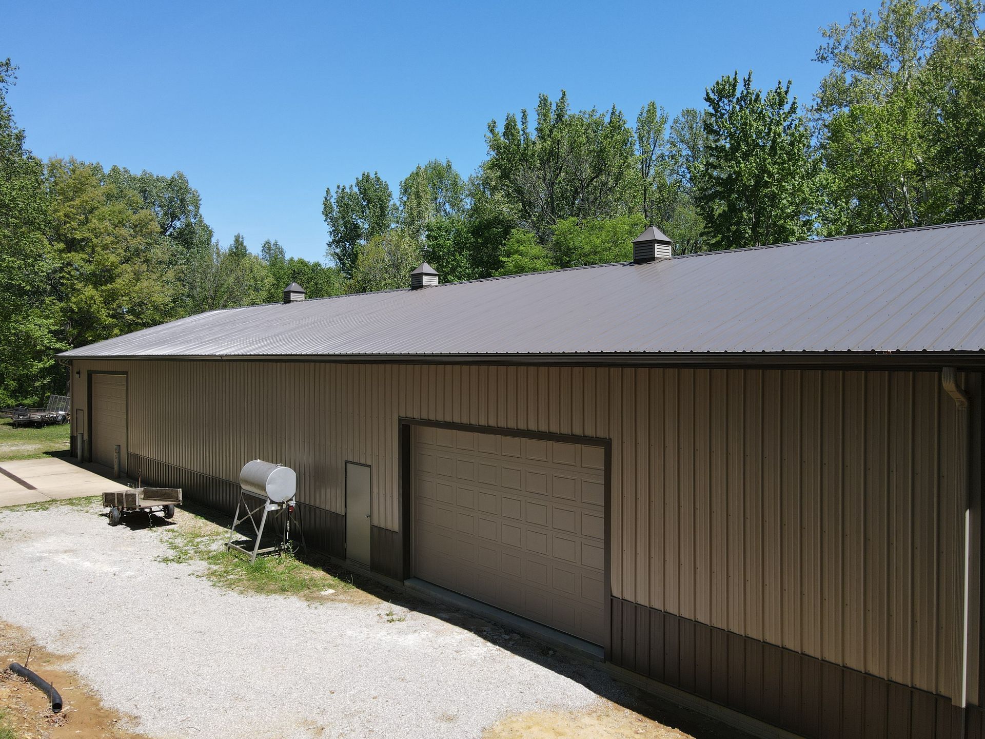 Long tan metal barn with gray roof and garage door, beside a gravel drive under blue sky