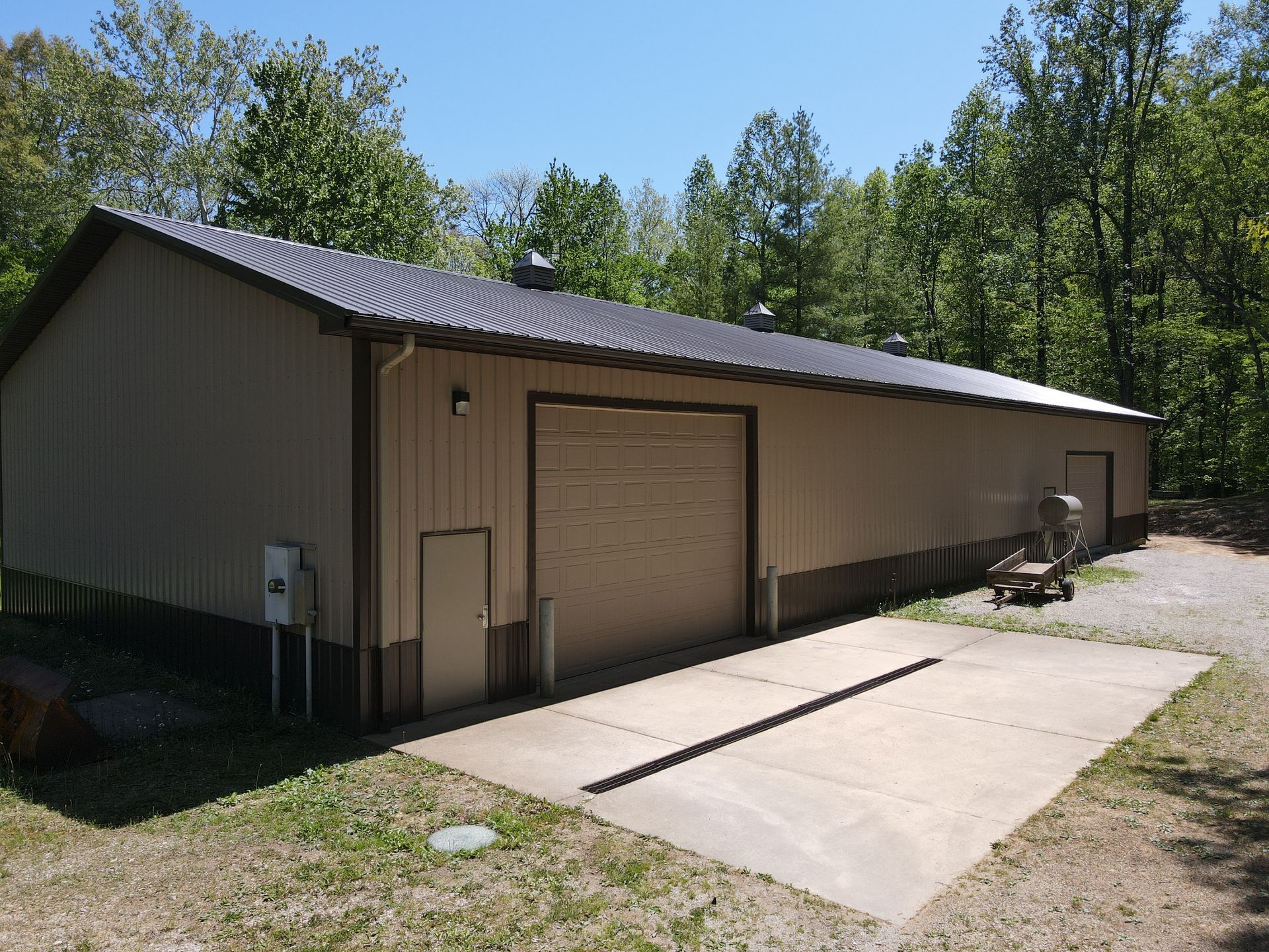 Tan metal garage with a roll-up door beside a concrete driveway and trees under a clear sky