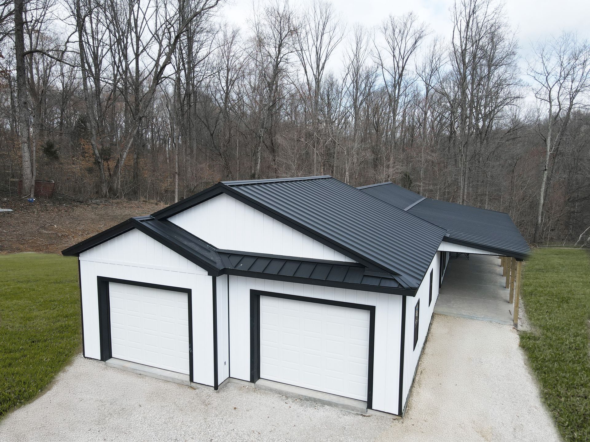 Detached white garage with dark roof and two doors, beside a driveway in a wooded area