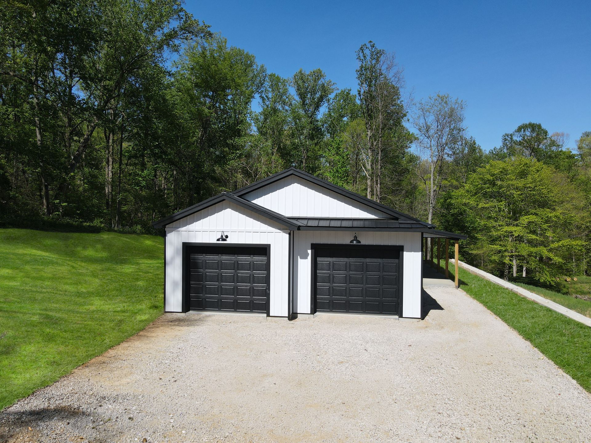 Detached two-car garage with black doors, white trim, and a gravel driveway surrounded by trees