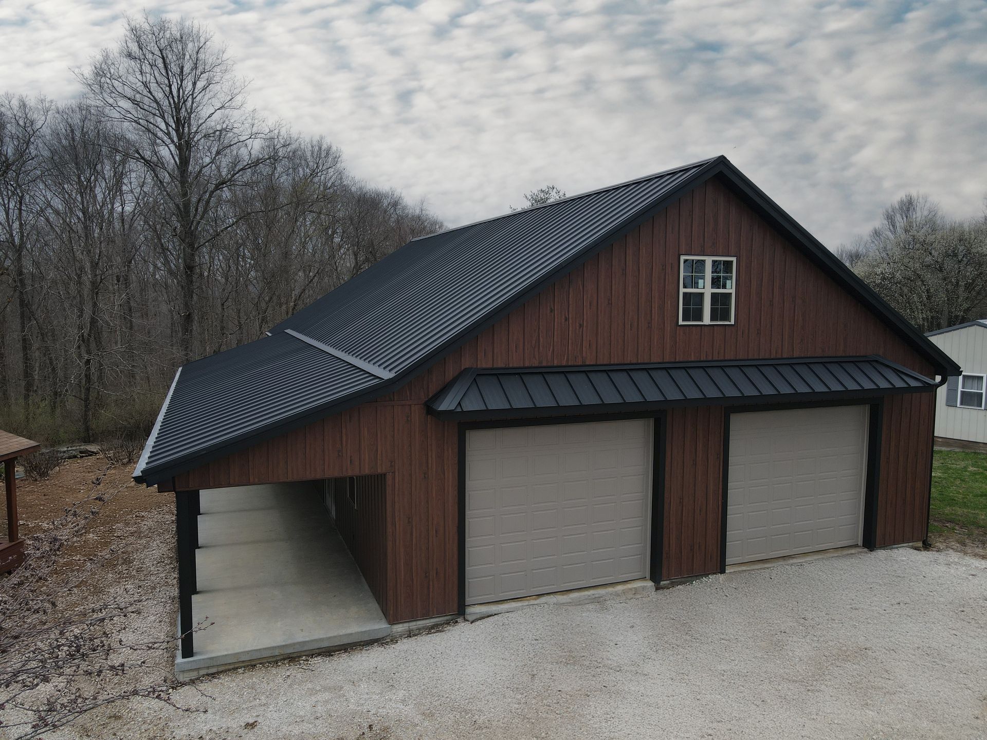 Brown garage with three gray doors and a dark roof on a gravel driveway, surrounded by bare trees.