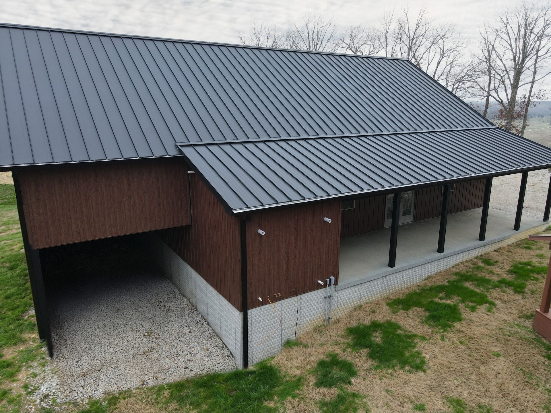 Brown barn with dark metal roof, open front bay, and gravel driveway on a grassy lot.