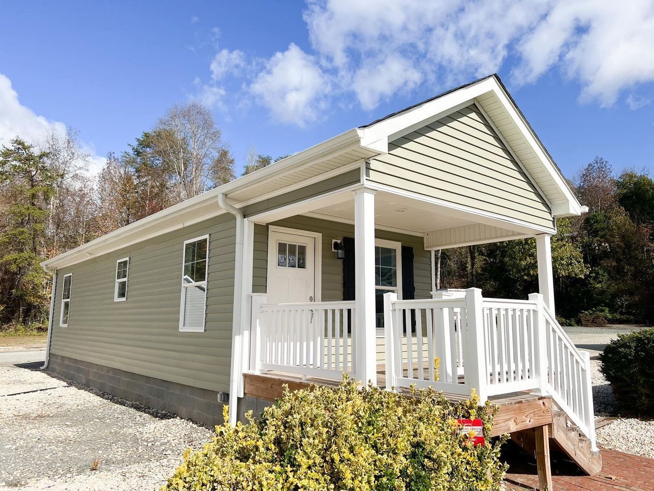 A sage green cottage with a white porch and railing, set on a gravel lot under a blue sky with scattered clouds.