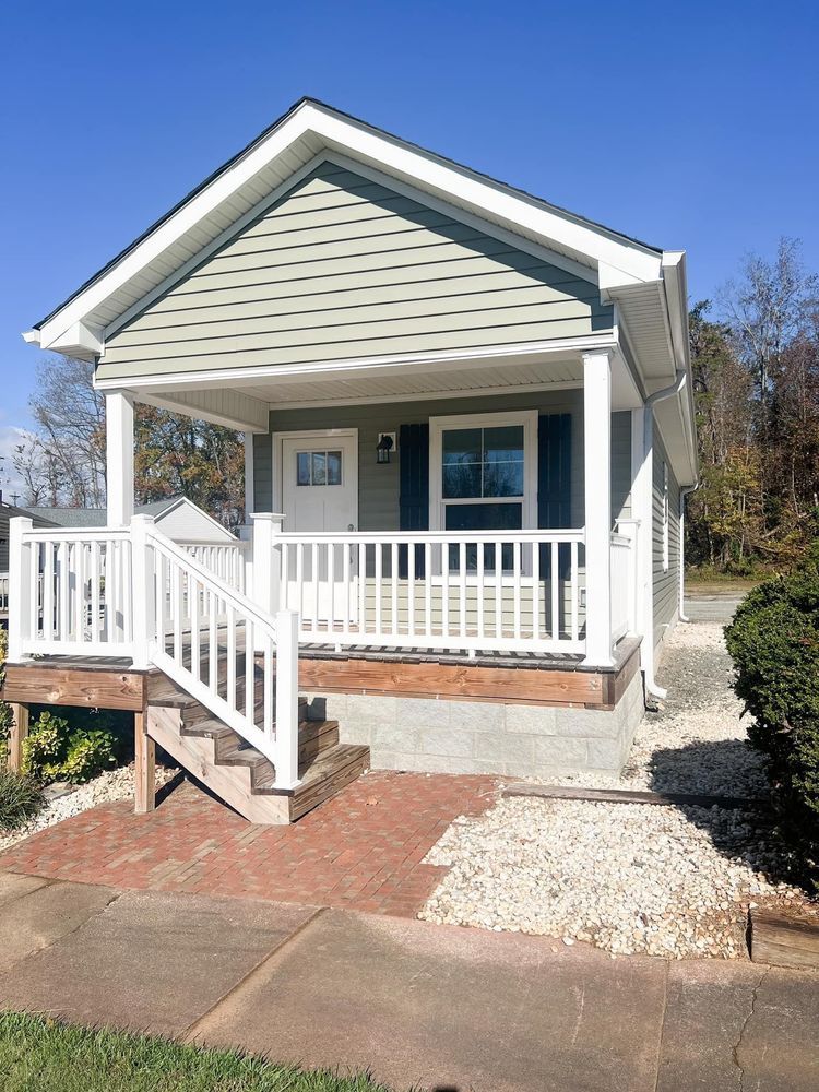 A small, light green cottage with a white front porch, stairs, and a red brick path, set against a clear blue sky.