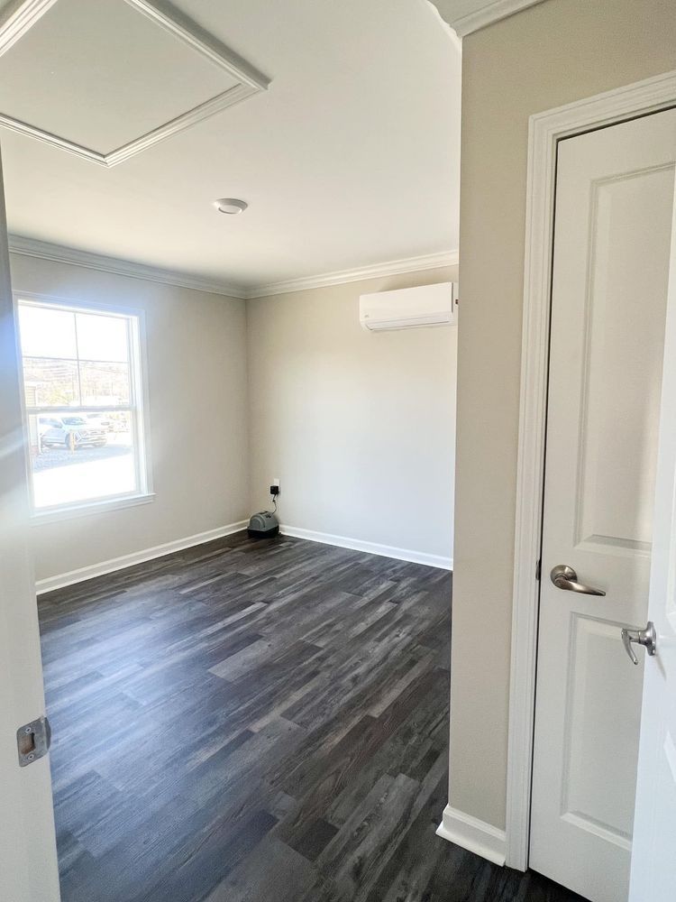 An empty bedroom with light-colored walls, dark wood-grain floors, a window, a ceiling attic hatch, and a white door.