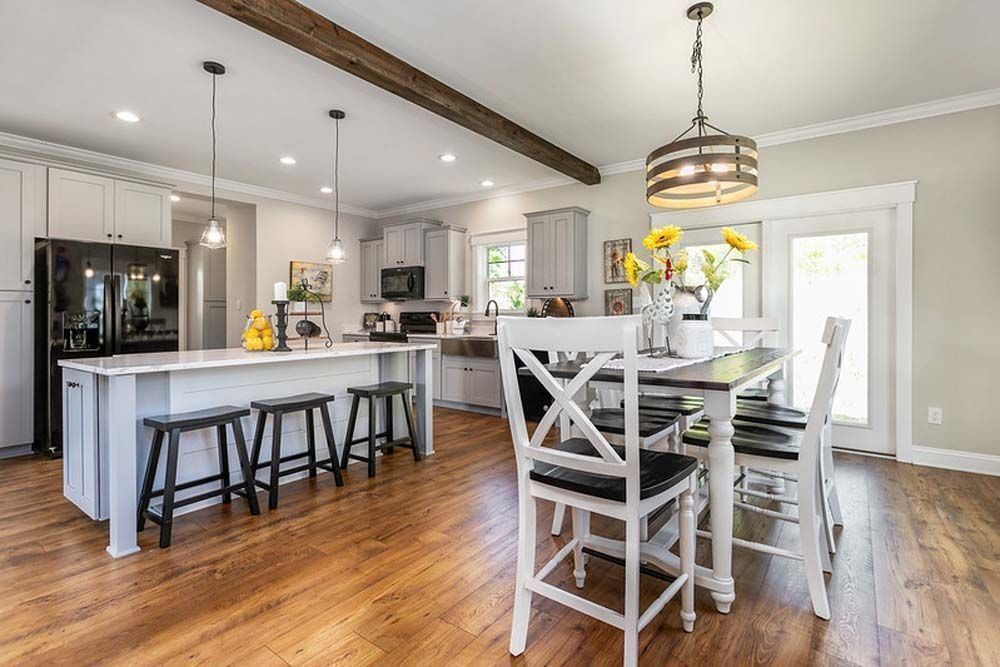 A modern kitchen and dining area featuring a white island with black stools, wood flooring, and farmhouse-style decor.