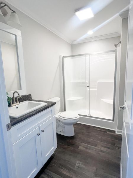 A bright, modern bathroom with a white vanity, a sink, a toilet, and a glass-enclosed shower on a dark wood-look floor.