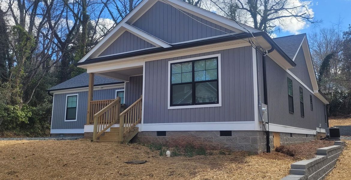 A newly constructed gray, single-story house with a wooden front porch stands on a sloped, grassy lot on a sunny day.