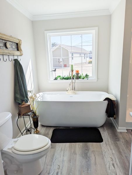 A freestanding white bathtub sits under a window in a bathroom with light grey walls and wood-look flooring.