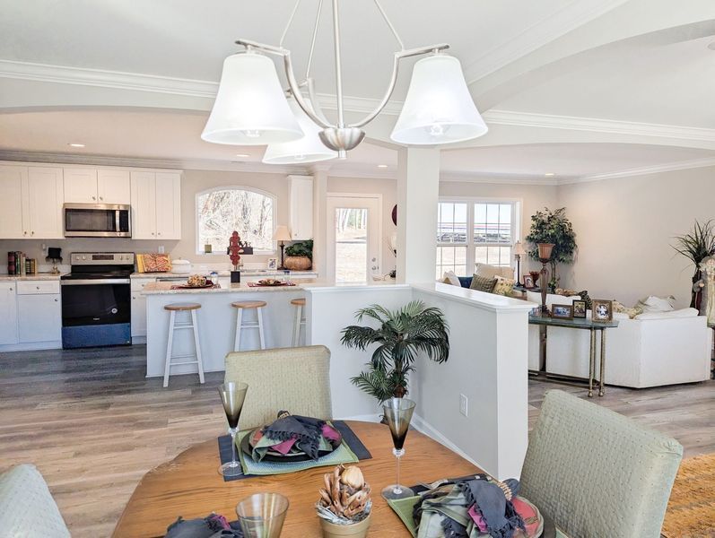 A dining table in the foreground opens into a bright kitchen and living area with light walls and wood-look flooring.