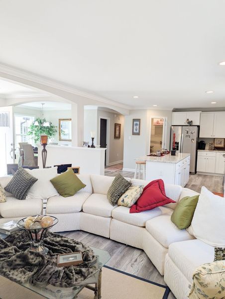 A cozy, white-walled living room with a large, cream-colored sectional sofa, colorful throw pillows, and an open kitchen.