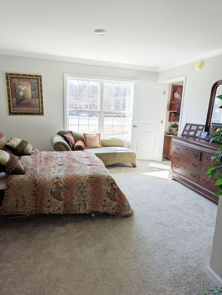 A bedroom featuring a bed with a patterned comforter, a chaise lounge by a window, and a wooden dresser with a mirror.
