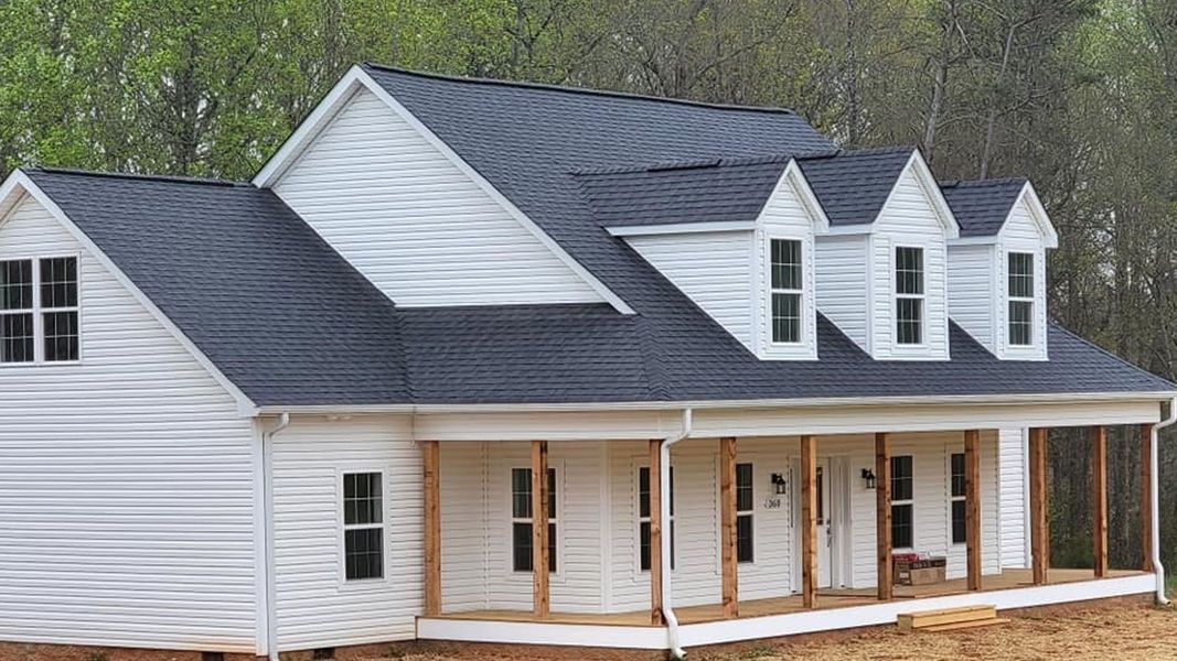 A two-story white house with a dark roof and a covered front porch, situated in a wooded area.