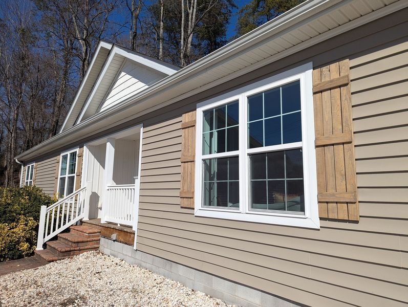 Tan, horizontal-sided house with white trim, a small porch, and wooden shutters on the windows against a wooded backdrop.