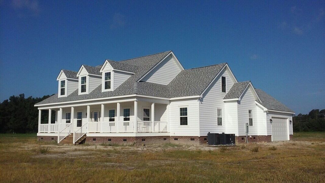A white farmhouse-style home under construction, featuring a porch, dormer windows, and a grey roof, set in a field.