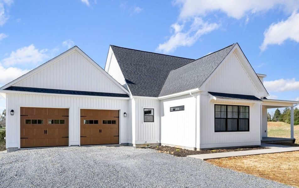A white modern farmhouse with vertical siding, black roof, and two brown garage doors under a blue sky.