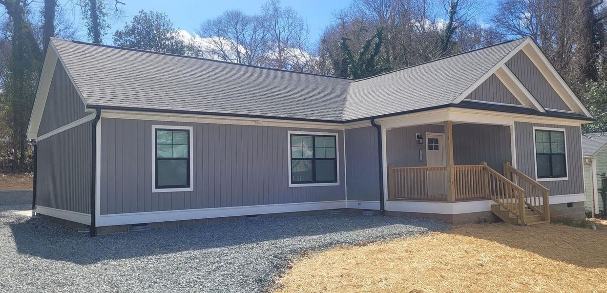 A single-story gray house with vertical siding, a metal roof, and a small front porch, surrounded by gravel and mulch.