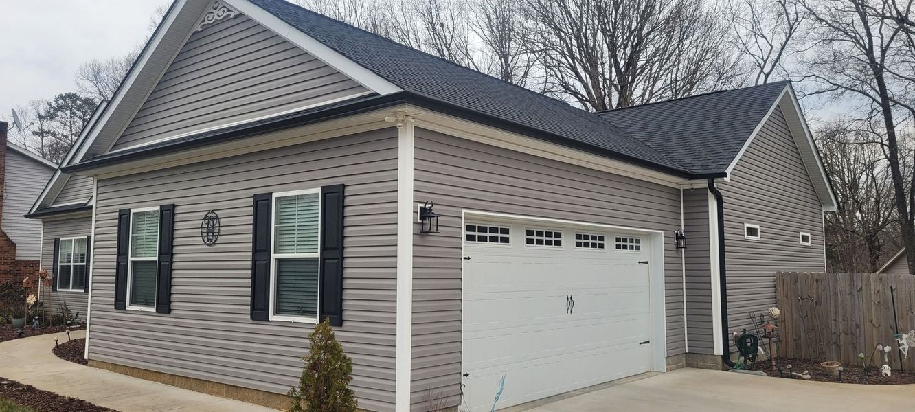 A single-story gray vinyl-sided house with a black metal roof, white garage door, and a concrete driveway.