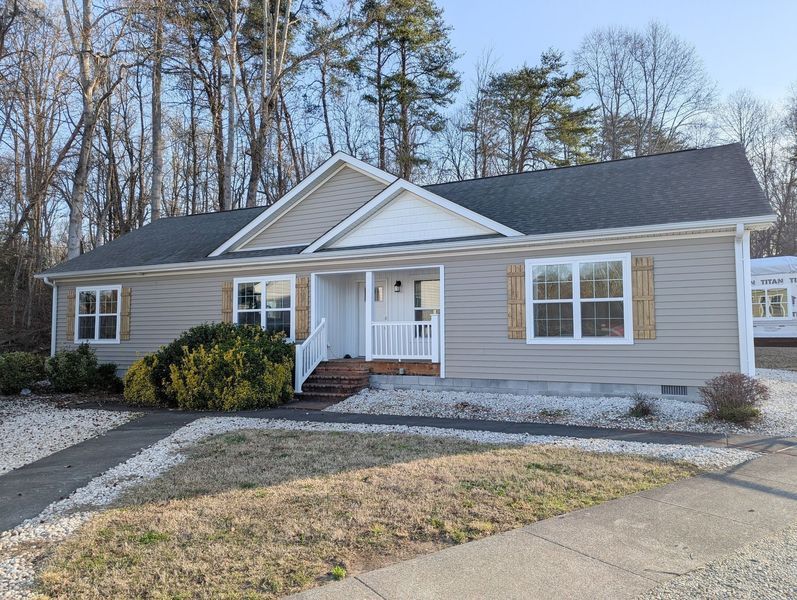 A light gray, single-story house with a white gabled porch, shuttered windows, and a gravel yard near trees.
