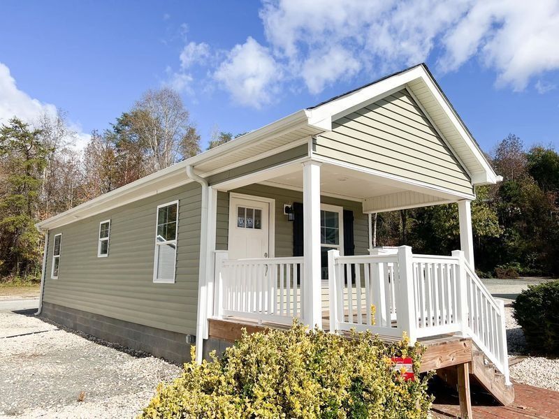 A small, sage-green cottage with white trim, a gabled porch, and white railing, set in a gravel lot under a blue sky.