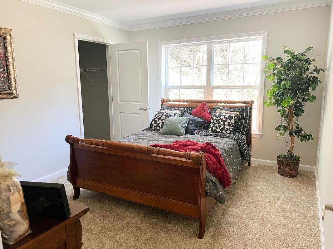 A bedroom with a wooden sleigh bed, patterned bedding with a red throw, a window, a closet door, and a potted tree.