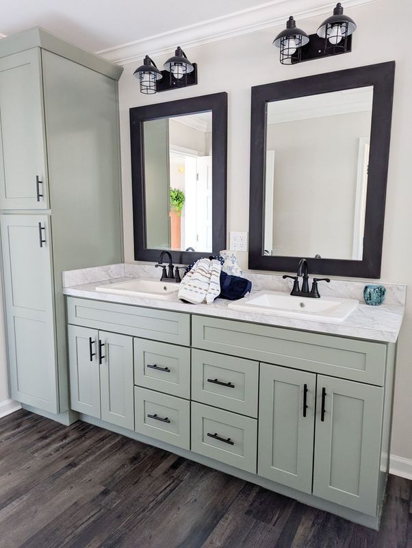 A double vanity with sage green cabinets, black hardware, white countertops, two sinks, and black-framed mirrors.
