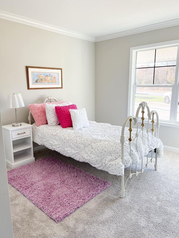 A bedroom with a white metal bed, polka-dot bedding, vibrant pink pillows, a bedside table, and a purple rug.