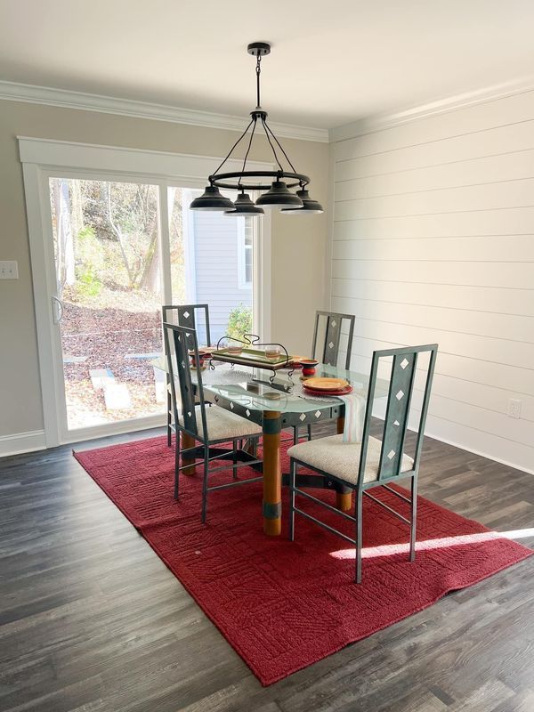 Dining area with a glass-topped table, four metal chairs on a red rug, and a rustic chandelier by a sliding glass door.