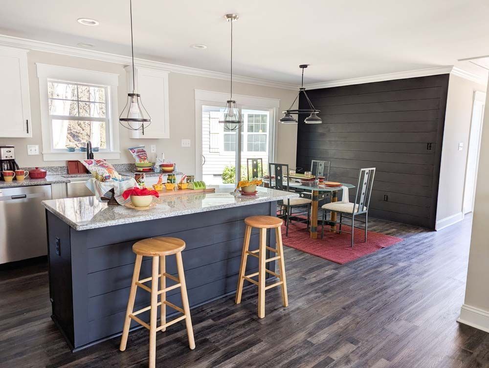 A kitchen with a dark blue island and two stools, white cabinets, and a dining area with a dark wood-paneled feature wall.