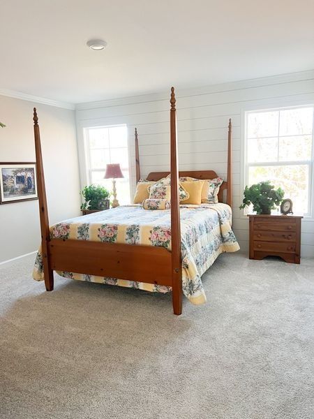 A four-poster wooden bed with floral bedding in a bright bedroom featuring shiplap walls, windows, and gray carpeting.