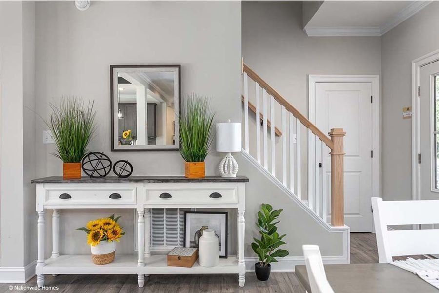 A white console table with decor, a mirror, and green plants sits in an entryway next to a staircase.