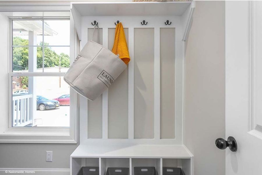 White mudroom bench with coat hooks, a hanging laundry bag, an orange cloth, and storage bins, located near a window.