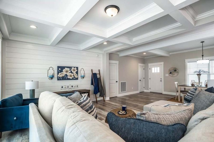 A bright living room with a beige sofa, dark blue armchair, shiplap wall, coffered ceiling, and entry door.
