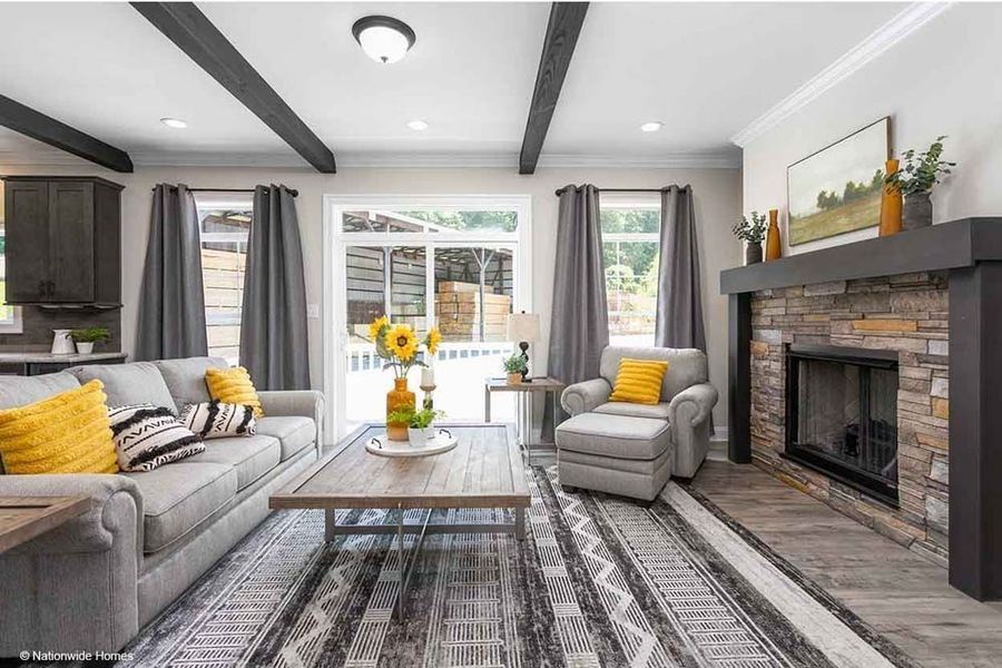 A cozy living room with a grey sofa, matching armchair, wood-top coffee table, and stone fireplace under dark ceiling beams.