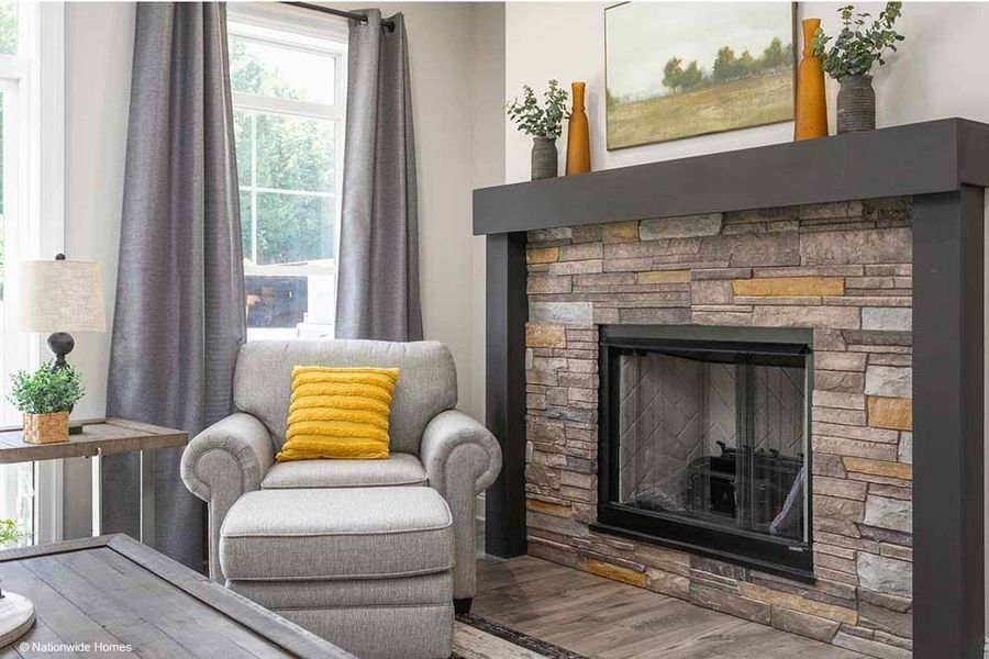 A gray armchair with a yellow pillow and matching ottoman sit beside a stone fireplace with a dark gray mantle.