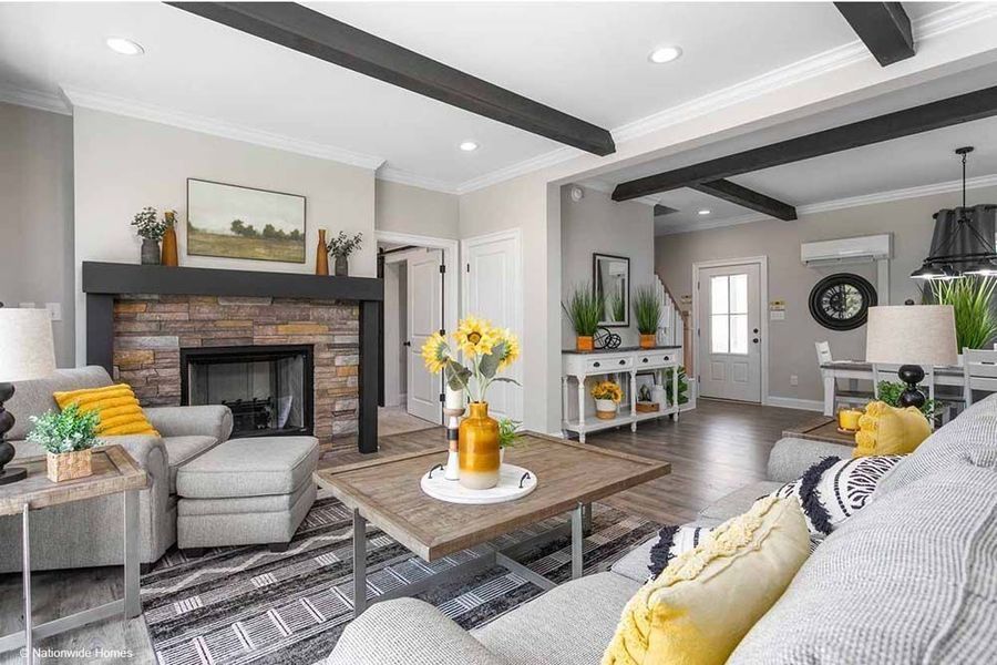A cozy living room with a stone fireplace, grey seating, yellow accents, dark ceiling beams, and a view of the entryway.