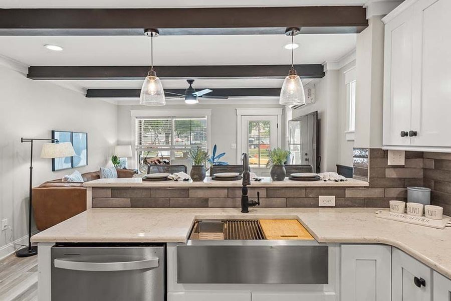 Modern kitchen with a stainless steel farmhouse sink and stone backsplash, looking toward a living area with exposed beams.