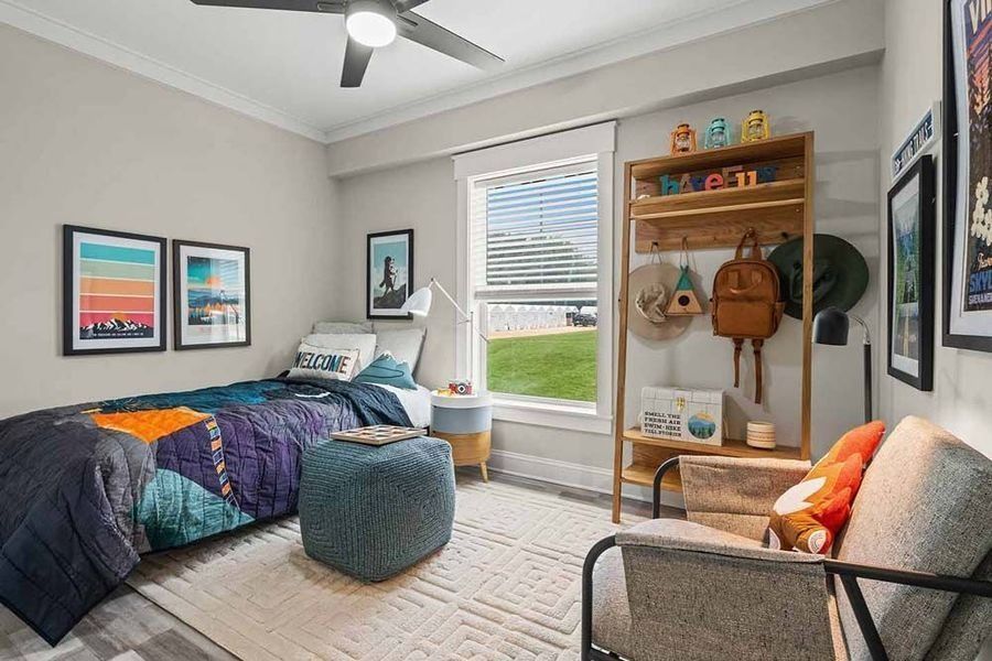 A cozy bedroom with a twin bed, a blue ottoman, a wooden open wardrobe, and a chair facing a window with a green view.