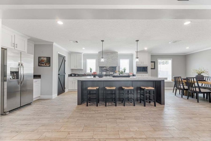 A bright, modern kitchen with white cabinets, a large dark island with four stools, stainless appliances, and tiled floors.
