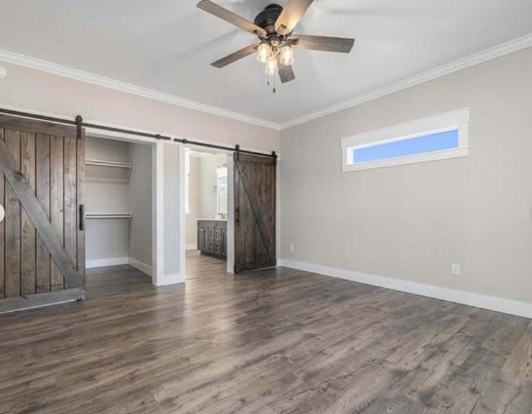 A modern bedroom with wood floors, light gray walls, a ceiling fan, and sliding barn doors leading to a closet and bath.
