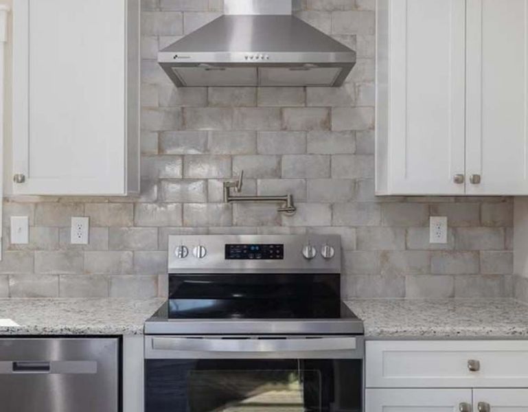 A kitchen stove and stainless steel range hood with a light-colored, rectangular tiled backsplash and white cabinets.