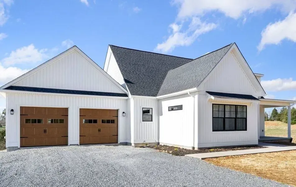 A new white farmhouse-style home with a two-car garage and grey roof under a blue, partly cloudy sky.