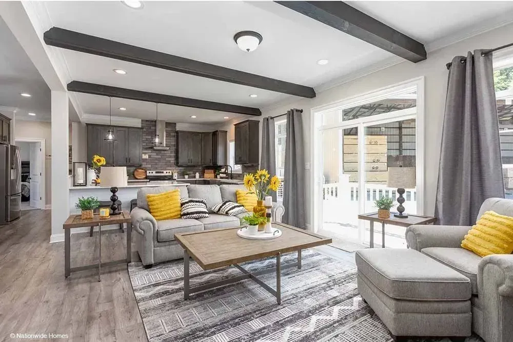 A bright living room features a grey couch, wooden coffee table, and an open kitchen with dark cabinets and ceiling beams.