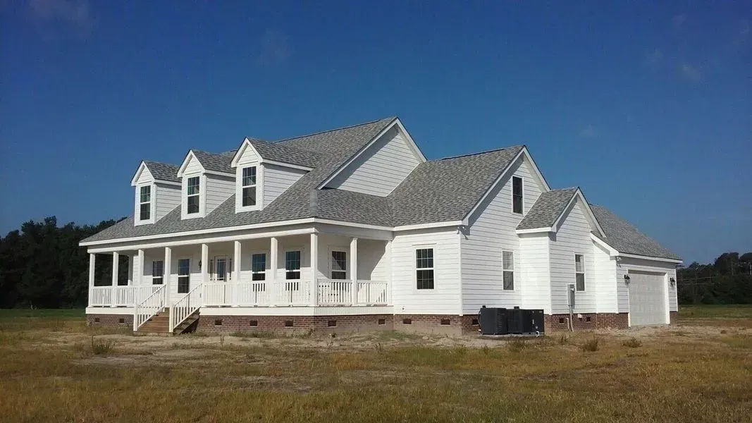 A white, two-story farmhouse with a porch and dormer windows, set in a dry, open field under a clear blue sky.