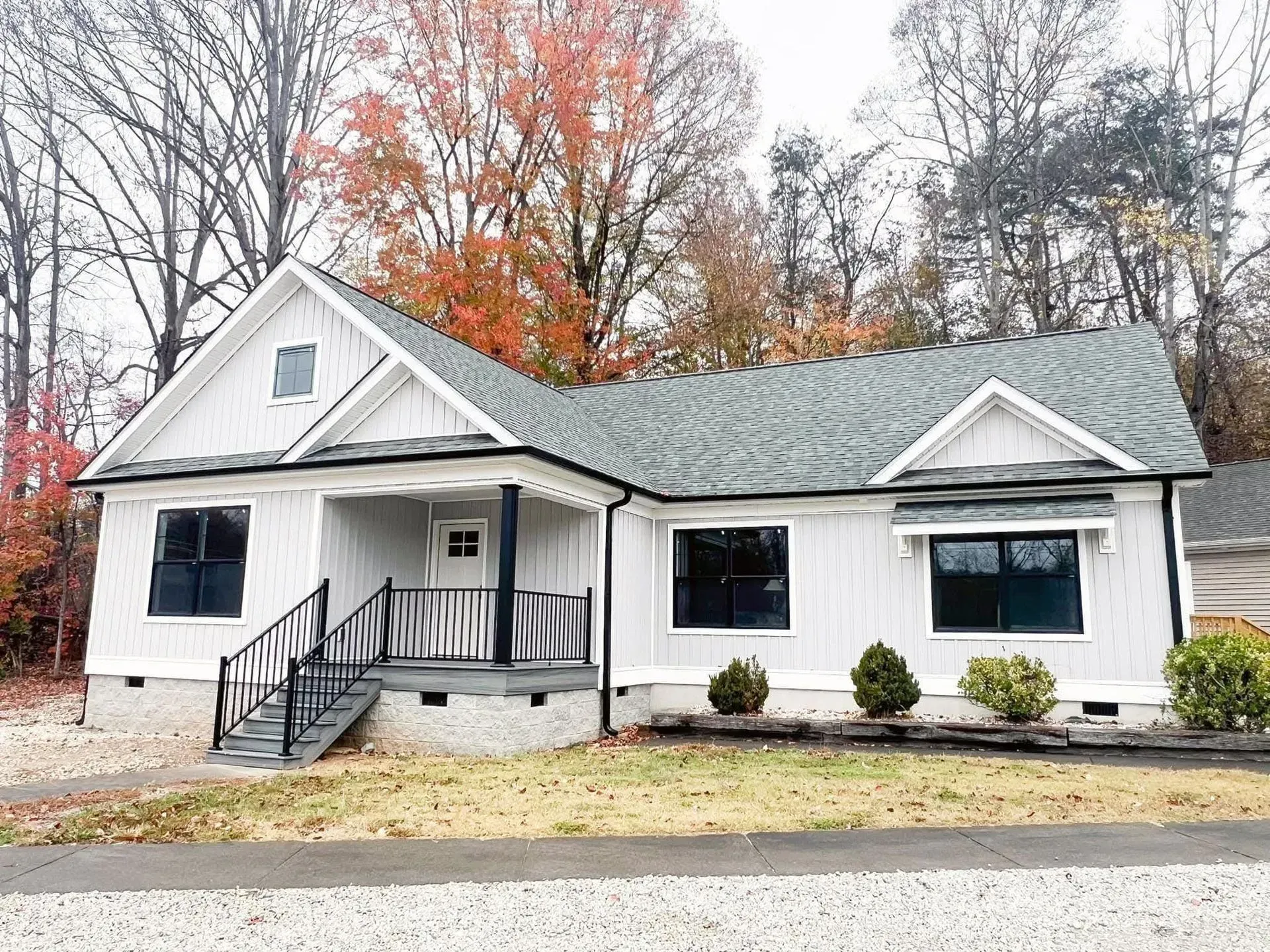 A white, one-story house with a grey shingled roof, black trim, and a small front porch, set against autumn trees.