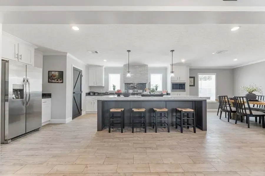 A spacious, modern kitchen featuring a dark island with four stools, stainless steel appliances, and beige tile floors.